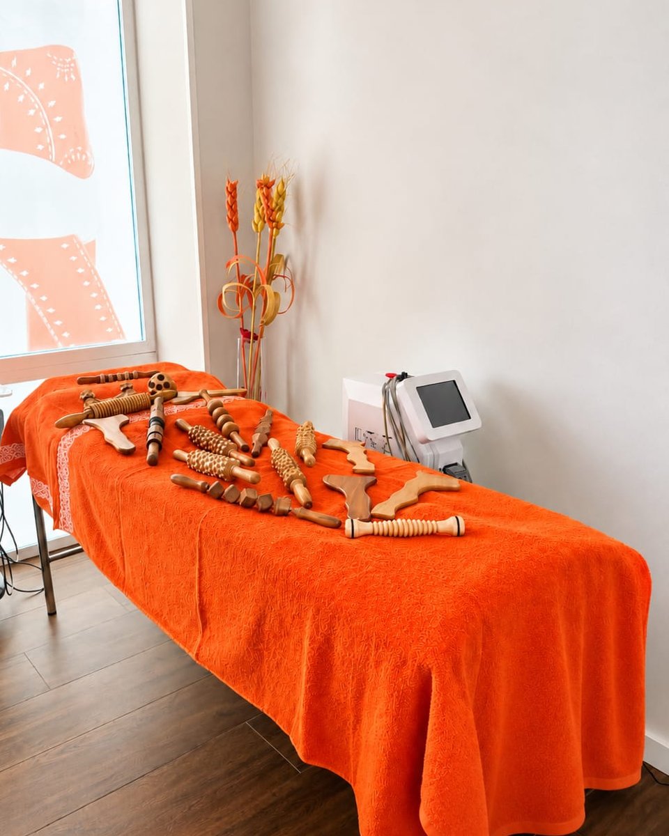 Cabina de Luz & Belleza: camilla con manta naranja y utensilios de madera para maderoterapia, paredes blancas, suelo de madera y luz natural
